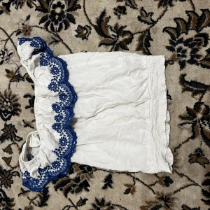 Vit offshoulder topp med blå spetsdetaljer - Säljer en söt vit offshoulder topp med vackra blå spetsdetaljer längs kanten. Perfekt för sommardagar eller en avslappnad fest. Toppen är i bra skick och har en luftig passform.