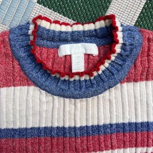 Randig stickad tröja i rött, blått och vitt - Säljer en färgglad stickad tröja med ränder i rött, blått och vitt. Tröjan har en ribbad struktur och trekvartsärmar. Den har en rund halsringning med volangdetaljer.