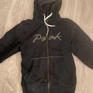 Svart hoodie från Peak Performance - Säljer en svart hoodie från Peak Performance med dragkedja och justerbar huva. Perfekt för kyliga dagar med sin bekväma passform och praktiska fickor framtill.