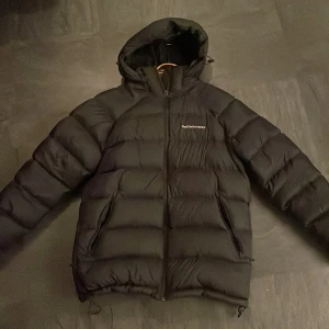 Svart dunjacka från Peak Performance - Säljer en svart dunjacka från Peak Performance i mycket bra skick. Jackan har en dragkedja framtill och är perfekt för kalla vinterdagar. Den är långärmad och har en skön huva för extra värme. Perfekt för både vardag och utomhusaktiviteter!