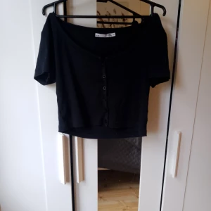 Svart kortärmad topp med knappar - Säljer en svart kortärmad topp med knappar framtill. Toppen är croppad och har en ribbad textur som ger en snygg look. Perfekt för både vardag och fest!