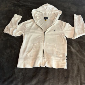 Vit hoodie från Ralph Lauren - Säljer en snygg och stilren vit hoodie från Ralph Lauren. Den har en dragkedja framtill och en klassisk broderad logga på bröstet. Perfekt för en avslappnad och bekväm stil. Passar både till vardags och lite finare tillfällen.