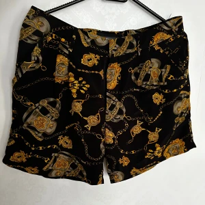 Svarta shorts med kedjemönster - Svarta shorts i strl38 med ett coolt mönster i guld. De har en normal passform och är perfekta för en somrig dag eller en festkväll. Materialet är tunn, mjuk och bekvämt. Perfekt för dig som vill sticka ut lite extra!