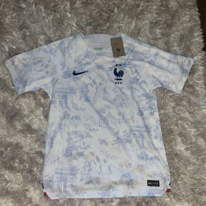 Vit fotbollströja från Nike - Säljer en vit fotbollströja från Nike med ett snyggt blått mönster. Tröjan har korta ärmar och är i nyskick, fortfarande i originalförpackning. Perfekt för fotbollsfans som vill ha något unikt och stilrent!