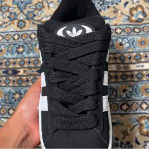 Säljer ett par svarta Adidas Campus sneakers i nyskick. Skorna har den klassiska tre-randiga designen i vitt och är gjorda i skinn. De kommer med extra vita skosnören. Perfekta för både vardag och fest! Aldrig Använda 