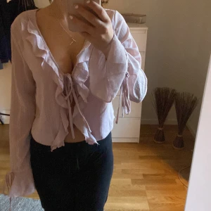 Ljusrosa blus med volangdetaljer - Säljer en superfin ljusrosa blus med volangdetaljer och knytning framtill. Blusen har långa ärmar. Nästan aldrig använd