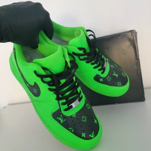 Custom LV Neon Green Nike Air Force 1 - Custom LV Neon Green Nike Air Force 1. Säljer ett par unika gröna Nike Air Force med svart mönster och texten 'LAST KING' på hälen. Skorna har en cool design med svarta snören och en svart Nike-logga. Perfekta för att sticka ut i mängden!