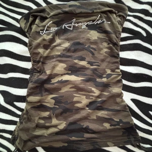 Camouflage tubtopp med strassdetaljer - Säljer en cool camouflage tubtopp med glittrande strassdetaljer på framsidan. Perfekt för en avslappnad men ändå stilfull look. Toppen är i ett mjukt och bekvämt material, idealisk för sommarens alla äventyr.