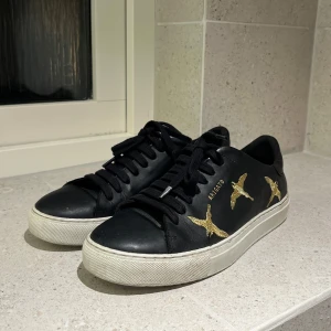 Svarta Axel Arigato skor - Säljer ett par svarta sneakers i skinn med snygga guldfärgade fågelbroderier på sidorna. Skorna har en vit sula och svarta snören. De är i bra skick och perfekta för både vardag och fest. Storlek 36 men passar mig som har 37. Dustbag finns!