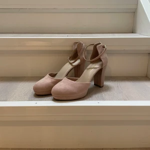 Rosa mocka pumps med klack - Säljer ett par superfina rosa pumps i ljusrosa. De har en elegant klack & en rem runt ankeln som ger extra stöd. Perfekta för fest eller bal! De är helt oanvänd och redo att användas. Lite damm på skorna då de legat i hyllan rätt länge. Nypris 349kr