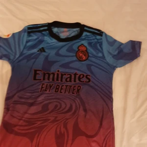 Blå och röd fotbollströja från Adidas - Snygg fotbollströja i blått och rött med svart mönster. Tröjan har korta ärmar och klubbmärke på bröstet. Texten 'Emirates Fly Better' pryder framsidan och 'VINI JR. 20' finns på baksidan.