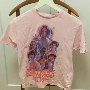 Rosa Stranger Things t-shirt från H&M - Cool rosa t-shirt från H&M med Stranger Things-tryck på framsidan. Perfekt för fans av serien! Kortärmad och tillverkad i mjuk bomull för en bekväm passform.