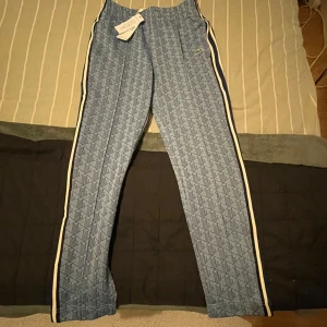 Blå mönstrade byxor från Lacoste - Snygga blå byxor från Lacoste med ett geometriskt mönster och vita sidoremsor. Byxorna har en avslappnad passform och är perfekta för en snygg look. Lacostes ikoniska krokodillogga pryder framsidan.