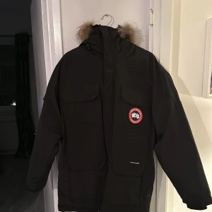 Svart dunjacka från Canada Goose - Säljer en svart dunjacka från Canada Goose med pälsdetaljer på huvan. Jackan har en klassisk design med dragkedja och flera praktiska fickor. Perfekt för kalla vinterdagar.