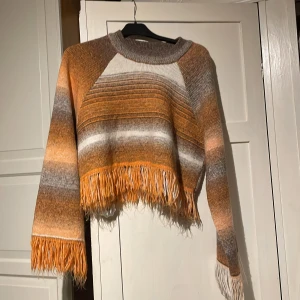 Randig tröja med fransar från Zara - Säljer en cool randig tröja från Zara i orange, grått och vitt. Tröjan är croppad med fransar vid nederkanten och ärmsluten, vilket ger den en unik look. Perfekt för höst och vår!