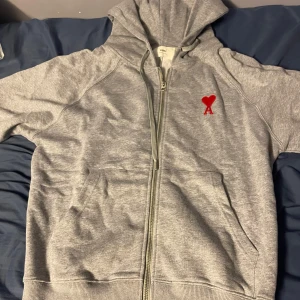 Ami Paris tröja zip - Säljer en stilren grå hoodie från AMI Paris med ett rött hjärta broderat på bröstet. Tröjan har dragkedja och huva, perfekt för en avslappnad look.