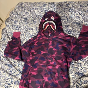 Camouflage hoodie från A Bathing Ape - Säljer en cool hoodie från A Bathing Ape med ett unikt lila och rosa camouflage-mönster. Hoodien har en dragkedja och ett hajmotiv på huvan. Den är dekorerad med bokstäverna WGM i olika färger på sidan.
