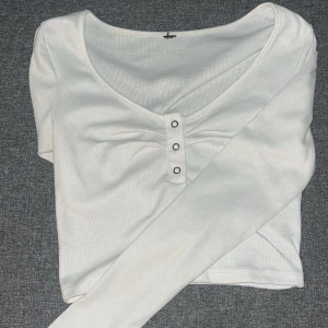 Vit ribbad långärmad topp - Säljer en stilren vit ribbad topp med långa ärmar och v-ringad design. Toppen har knappar framtill som en snygg detalj.