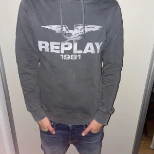 Replay hoodie - Snygg grå hoodie från Replay med tryck '1981' och en cool örn på bröstet. Perfekt för en avslappnad stil. Den har en justerbar huva med snören och ribbade muddar vid ärmslut och nederkant.