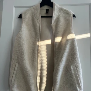  fleeceväst - Säljer en mysig vit fleeceväst med dragkedja. Perfekt för kyliga dagar och enkel att matcha med andra plagg. Västen har två fickor framtill.