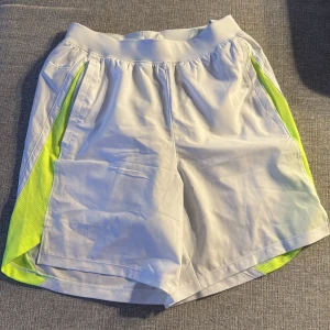 Vita träningsshorts med neongula detaljer - Snygga vita träningsshorts från Under Armour med neongula sidopaneler. Perfekta för träning med elastisk midja och snörning för bästa passform. Tillverkade i lätt och bekvämt syntetmaterial.