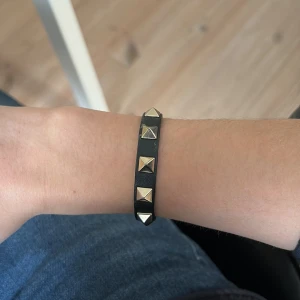 Svart armband med nitar från Valentino - Snyggt svart armband från Valentino med guldfärgade nitar och ett justerbart spänne. Perfekt accessoar för att ge en edgy touch till din outfit. Kommer med en praktisk förvaringspåse.