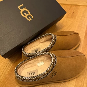 Ugg tasman - Mysiga bruna tofflor från UGG med mjukt foder och dekorativ kant. Perfekta för att hålla fötterna varma under kyliga dagar. Tofflorna har en bekväm passform och är tillverkade i mocka. Passar både inne och ute. Dem är endast använda 1 gång, fick i julklapp detta året därav priset. Slutsåld på de flesta ställen