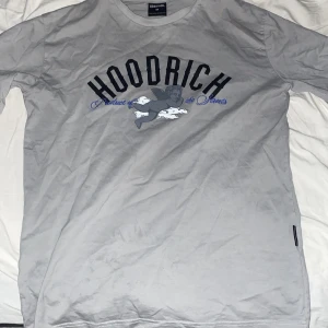Grå t-shirt från Hoodrich - Snygg grå t-shirt från Hoodrich med tryck fram och bak. Framsidan har texten 'Hoodrich' och en grafisk design. Baksidan pryds av ett stort 'H' och texten 'From Nothing To Something'. Perfekt för en avslappnad stil.