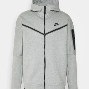 Grå hoodie från Nike - Snygg grå hoodie från Nike med dragkedja och huva. Jackan har en modern design med svarta detaljer och en praktisk ficka på ärmen. Perfekt för en sportig look. Dragkedjan går inte att stänga, men det går enkelt att laga