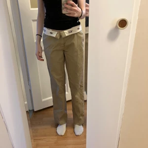 Beige byxor från Dickies - Säljer ett par klassiska beige byxor från Dickies med vit logotypdetalj på insidan av midjan. Byxorna har en rak passform och är perfekta för en avslappnad stil. De har en stilren design med knapp och dragkedja framtill.