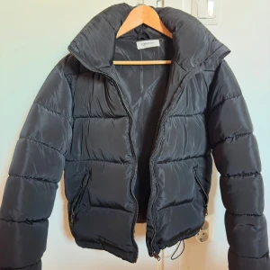 Svart pufferjacka - Snygg svart pufferjacka  med dragkedja och hög krage. Perfekt för kyliga dagar med sin vadderade design. Jackan har två praktiska fickor med dragkedjor. Storlek 36. Behöver tvättas då det finns lite smink på kragen.   Kan skickas om köparen står för frakten. 