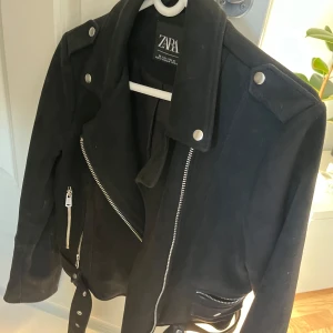 Svart skinnjacka från Zara - Snygg svart skinnjacka från Zara med dragkedjor och bälte i midjan. Jackan har långa ärmar och axelklaffar med knappar. Perfekt för en cool look.