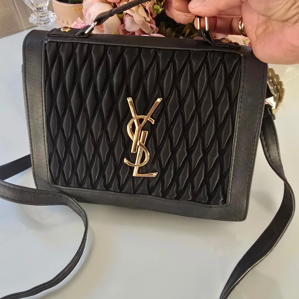 Snygg svart axelväska med quiltad design och guldfärgad logotyp på framsidan. Väskan har en justerbar axelrem och ett handtag för enkel bärning. Perfekt för att ge en elegant touch till din outfit. Inte äkta!. Laukut & Käsilaukut.
