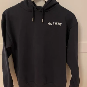 Mörkblå hoodie med text från Glamorous - Säljer en mörkblå hoodie från Gina Tricot med texten 'MA CHÉRIE' i vitt på framsidan och glittriga detaljer på baksidan. Tröjan har en klassisk huva med dragsko och långa ärmar. Perfekt för en avslappnad stil.