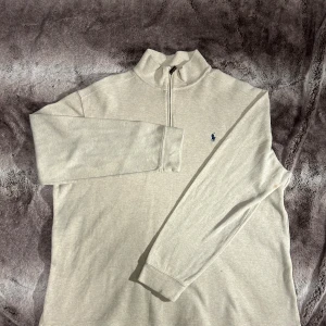 Ralph Lauren - Säljer en stilren beige tröja från Ralph Lauren med dragkedja och broderad logga på bröstet. Tröjan har långa ärmar och en hög krage, perfekt för kyligare dagar. Endast kemtvätt.