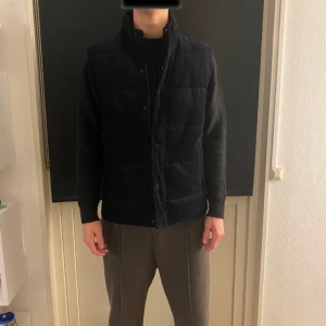 Svart quiltad väst från Dressmann - Säljer en snygg svart quiltad väst från Dressmann med dragkedja och knappar. Perfekt för lager-på-lager under kyliga dagar. Västen har en stilren design och är tillverkad i ett mjukt material. 