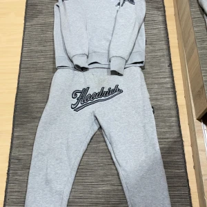 Grå hoodie och byxor från Hoodrich - Säljer ett set med grå hoodie och matchande byxor från Hoodrich. Hoodien har långa ärmar och en tryckt logga på bröstet. Byxorna har en elastisk midja och logga på benet. Perfekt för en avslappnad stil.