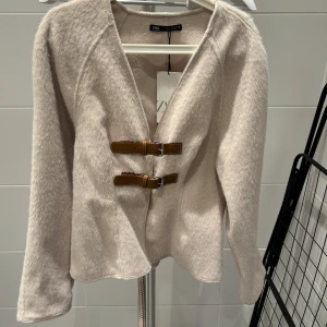 Beige jacka/blazer från Zara med bältesdetaljer - Snygg beige jacka från Zara med två bruna bältesdetaljer framtill. Jackan har en mjuk och fluffig textur och långa ärmar. Perfekt för en stilren look.