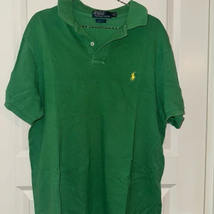 Grön pikétröja från Ralph Lauren - Säljer en klassisk grön pikétröja från Ralph Lauren med gul broderad logga på bröstet. Tröjan har korta ärmar och en stilren krage. Perfekt för en avslappnad men stilfull look.