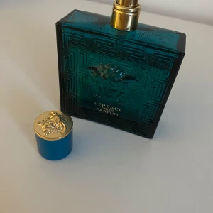 Versace Eros Parfum 85/100ml - Säljer en elegant Versace Eros Parfum i en stilren, fyrkantig flaska av mörkgrönt glas med ett detaljerat mönster. Flaskan har en gyllene spraypump och ett blått lock med gulddetaljer. Perfekt för den som vill ha en lyxig doftupplevelse. 85ml kvar ungefär. Kommer i ordinarie kartong.