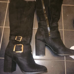 Svarta stövlar med klack och spännen - Snygga svarta stövlar i mocka med hög klack och dekorativa spännen. De har en praktisk dragkedja på sidan för enkel på- och avtagning. Perfekta för höst och vinter.