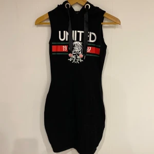 Svart ärmlös klänning från Nevyorker - Snygg svart ärmlös klänning från Newyorker med huva och tryck framtill. Klänningen har texten 'UNITED' och ett coolt mönster med ränder i rött och grönt. Perfekt för en avslappnad stil.