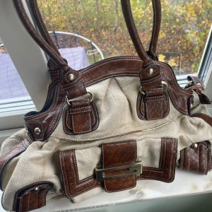 Beige och brun axelväska Guess  - Snygg beige axelväska med bruna skinn detaljer och metallspänne. Väskan har dubbla handtag och flera dekorativa remmar. Perfekt för en stilren look.