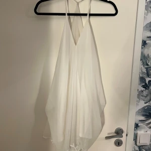 Vit klänning med tunna axelband  - Elegant vit klänning med ett flödande tyg, smala axelband och sjukt fin rygg detalj då axelbanden sträcker sig till ryggen. Perfekt för sommarens festligheter eller en stilren look. Klänningen har en luftig passform och är tillverkad i ett lätt material. Ganska kort för folk över 168. Kan rekommenderas som strand klänning eller som en lätt topp för folk som är långa. 