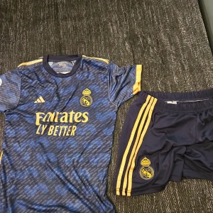 Blå Real Madrid fotbollströja från Adidas - Säljer en blå Real Madrid fotbollströja från Adidas med guldiga detaljer och korta ärmar. Tröjan har ett mönster i olika blå nyanser och klubbens emblem på bröstet. 
