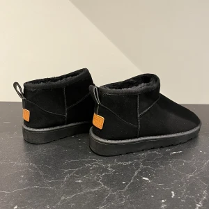 Svarta skor med foder - Snygga svarta boots med mjukt foder och en robust sula. Perfekta för kyligare dagar. De har en praktisk dragflik baktill och en dekorativ läderdetalj med logotyp. Passar bra för både stad och natur. Svarta oanvända skor. Säljs så storleken inte stämmer. På skon står det storlek 41 men det stämmer inte. Innermåttet är 24/24,5 cm. (Liknar uggs) skulle säga att skon är 38/39