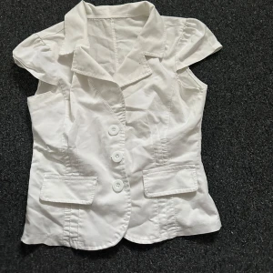 Vit blus med knappar - Säljer en stilren vit blus med korta ärmar och tre knappar framtill. Blusen har en klassisk krage och två dekorativa fickor. Perfekt för en elegant look och passar till fest!