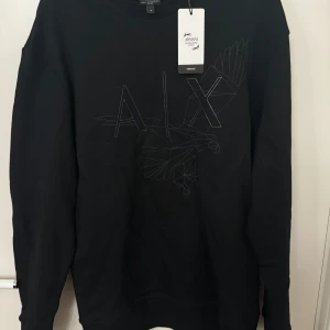 Svart tröja  Armani Exchange strl M - Snygg svart tröja från Armani Exchange med ett diskret mönster av en fågel och bokstäverna 'A|X' på framsidan. Tröjan har långa ärmar och en klassisk passform. Perfekt för en stilren look. Oanvänd med väldigt bra passform