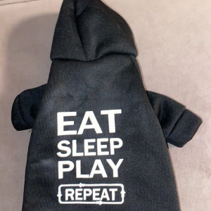 Svart hoodie med text till hundar  - Säljer en svart hoodie med texten 'EAT SLEEP PLAY REPEAT' i vitt. Tröjan har korta ärmar och en klassisk huva. I storlek S. aldrig använd💕