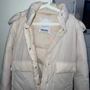  Dunjacka från Kenzo - Säljer en snygg krämvit dunjacka från Kenzo med huva och dragkedja. Jackan har en broderad detalj på fickan och är perfekt för kyliga dagar. Den är i storlek M och har en normal passform. Ord. pris runt 7000:- använd 2 gånger. 